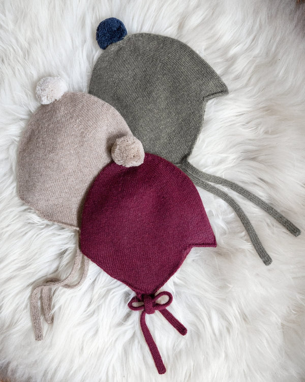 Three knitted baby bonnets in stone, green, and red on a fluffy white surface.