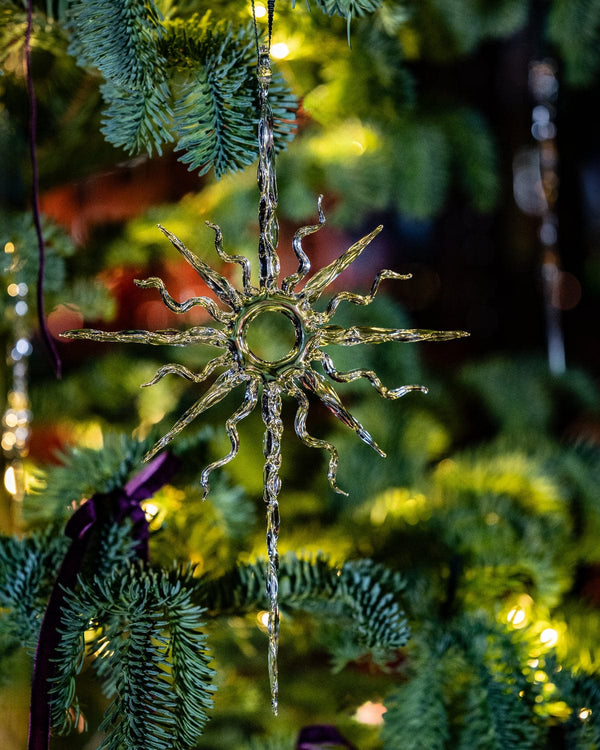 Decorative sun-shaped ornament hanging on a Christmas tree with greenery and lights in the background.