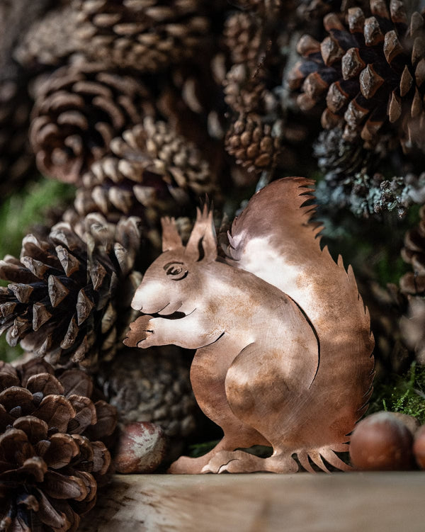 Copper squirrel figurine surrounded by pine cones and nuts on a wooden surface