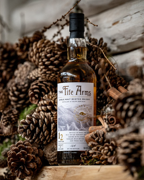 Bottle of 'The Fife Arms' whisky surrounded by pine cones on a wood plank.