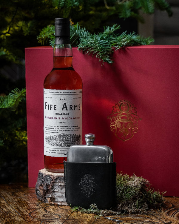 Bottle of 'The Fife Arms' whisky with a hip flask on a wooden surface with a red box and greenery in the background.