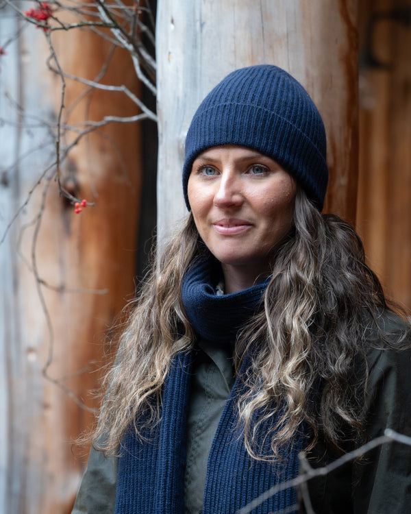 Woman wearing a blue beanie and scarf standing between two wooden posts with bare branches.