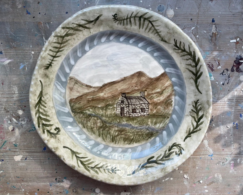 Ceramic plate with a landscape design featuring a cabin and mountains on a textured surface.