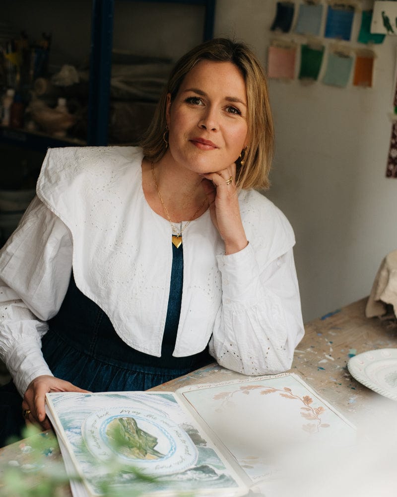 Woman sitting at a table with art supplies, wearing a white blouse.
